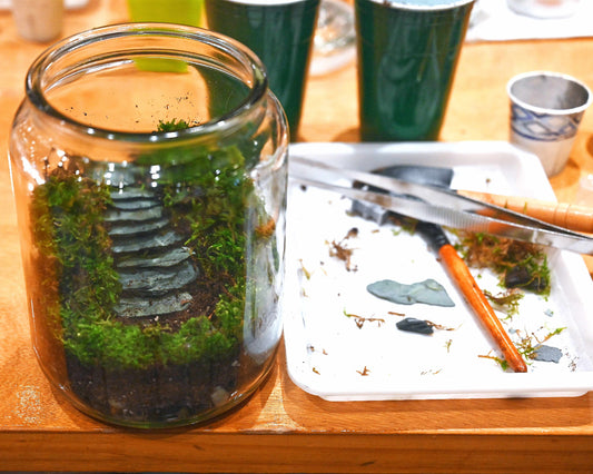 Glass jar with greenery on a wooden table with gardening tools and cups in the background.