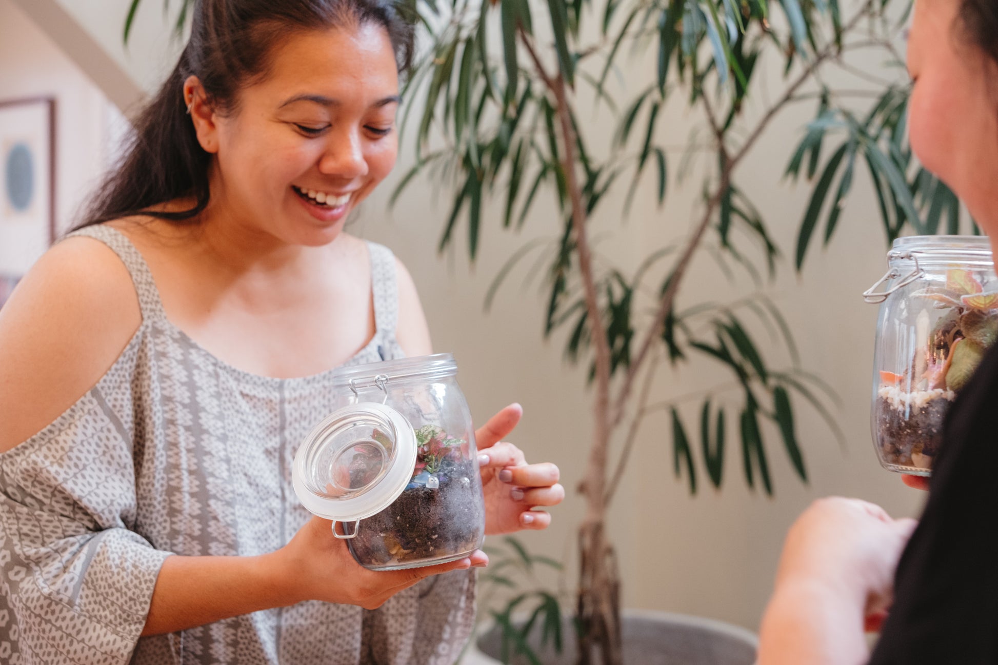 Two people interacting with a glass jar containing a plant, with a plant in the background.