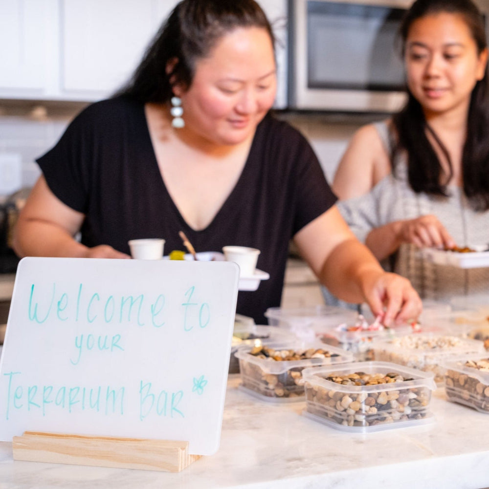 Two women in a kitchen with a sign that says 'Welcome to your Terrarium Bar' on a counter with containers of items.
