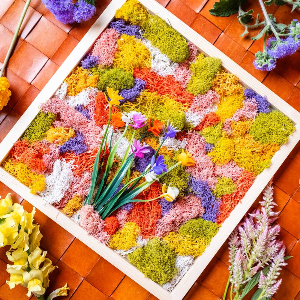 Colorful floral arrangement in a wooden frame on a woven surface with additional flowers around.