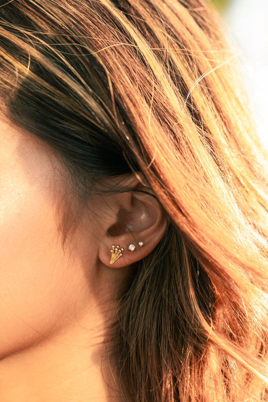 A close-up of a person's ear wearing a small gold geometric stud earring and a small silver stud, with warm sunlight filtering through brown and blonde hair.