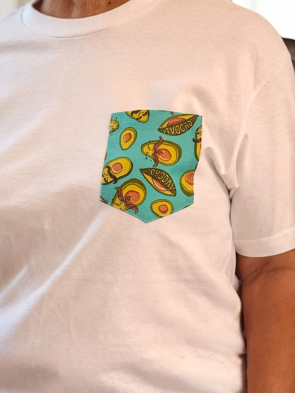 White t-shirt with a colorful avocado pattern pocket on a plain background