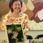 Woman holding a framed moss art piece with a colorful abstract painting in the background