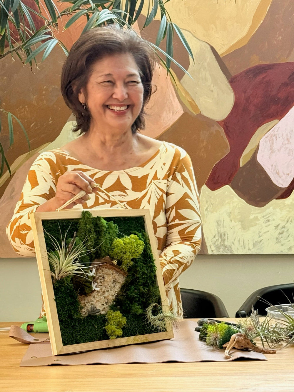 Woman holding a framed moss art piece with a colorful abstract painting in the background