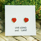 A pair of small red heart-shaped stud earrings displayed on a white card that reads 'LIVE LONG and PLANT', placed on a woven mat with green moss in the background.