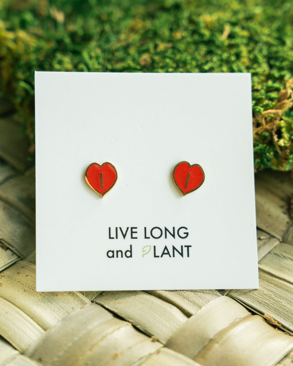 A pair of small red heart-shaped stud earrings displayed on a white card that reads 'LIVE LONG and PLANT', placed on a woven mat with green moss in the background.