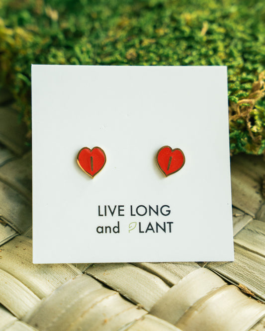 A pair of small red heart-shaped stud earrings displayed on a white card that reads 'LIVE LONG and PLANT', placed on a woven mat with green moss in the background.
