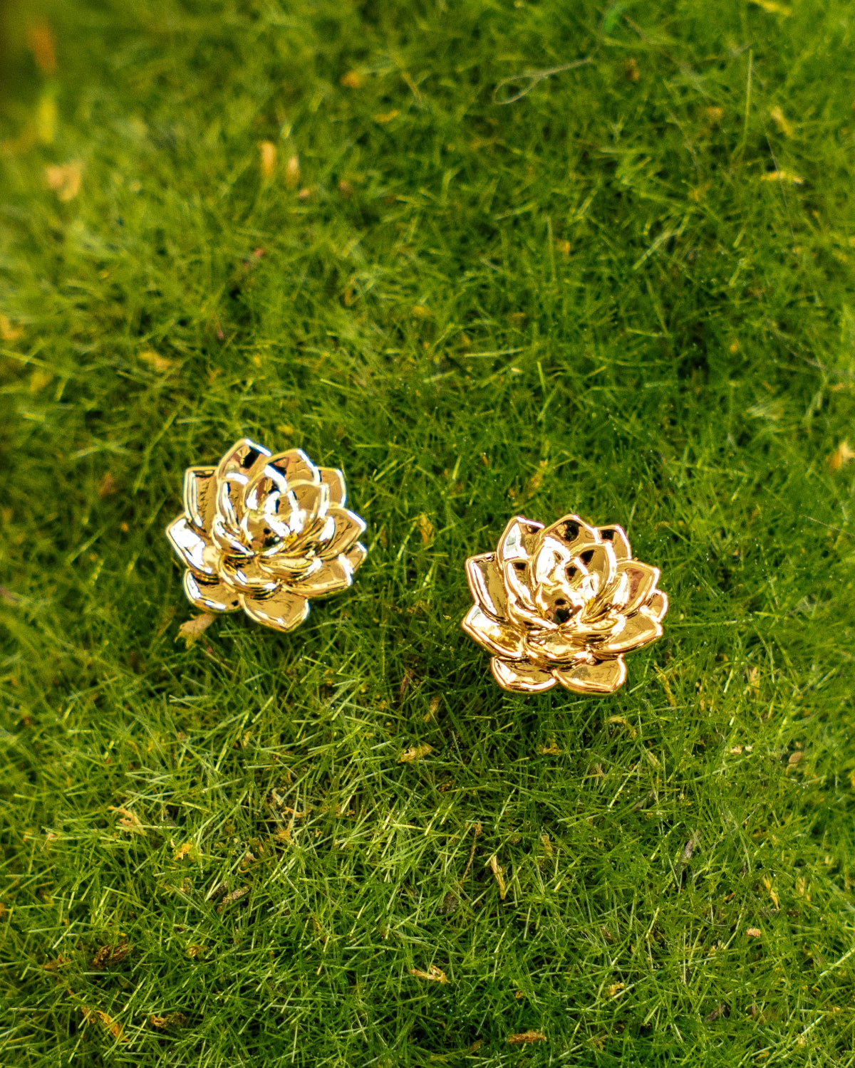 A pair of gold-toned flower stud earrings resting on a bed of green moss.