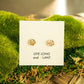 A pair of small gold floral stud earrings displayed on a white card that reads 'LIVE LONG and PLANT,' resting on green moss over a woven wooden surface.