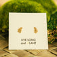 A pair of small gold fern-shaped stud earrings displayed on a white card that reads 'LIVE LONG and PLANT', set against a blurred green moss and woven natural background.