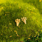 A pair of small gold stud earrings shaped like tiny mushroom clusters, resting on bright green moss with a soft blurred natural background.