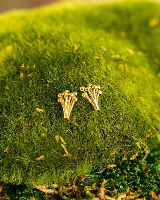 A pair of small gold stud earrings shaped like tiny mushroom clusters, resting on bright green moss with a soft blurred natural background.