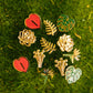 A set of nine botanical and heart-shaped enamel earrings arranged on a textured green moss background. The pins feature gold-plated metal outlines with red, green, and white enamel details, including hearts, roses, leaves, and monstera leaves.