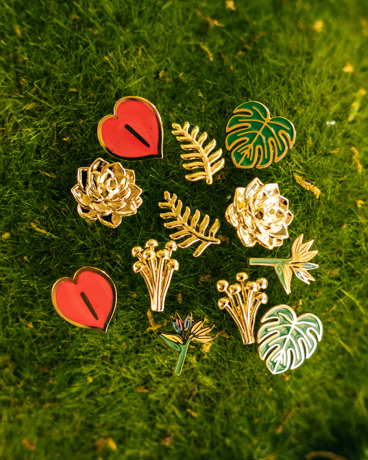A set of nine botanical and heart-shaped enamel pins arranged on a textured green moss background. The pins feature gold-plated metal outlines with red, green, and white enamel details, including hearts, roses, leaves, and monstera leaves.