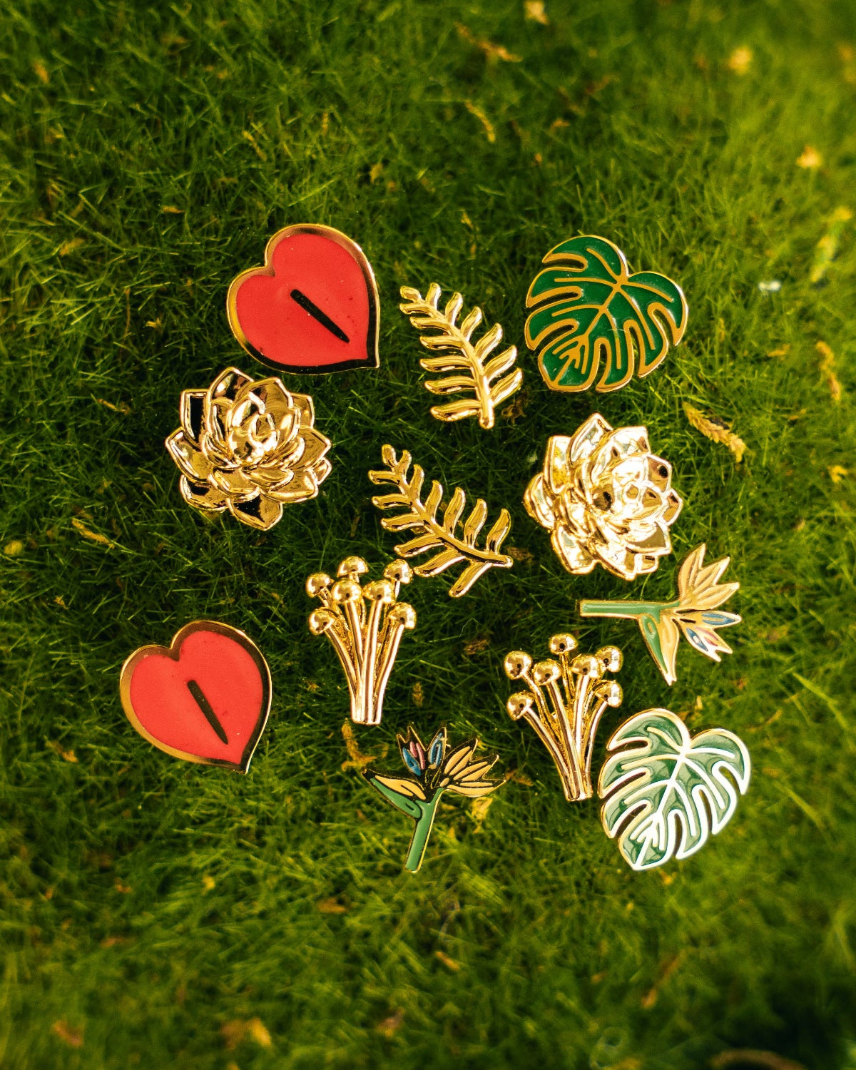 A set of nine botanical and heart-shaped enamel stud earrings arranged on a textured green moss background. The earrings feature gold-plated metal outlines with red, green, and white enamel details, including hearts, roses, leaves, and monstera leaves.