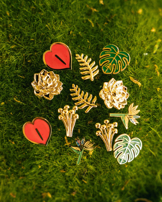 A set of nine botanical and heart-shaped enamel stud earrings arranged on a textured green moss background. The earrings feature gold-plated metal outlines with red, green, and white enamel details, including hearts, roses, leaves, and monstera leaves.