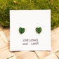 A pair of green monstera leaf stud earrings displayed on a white card that reads 'LIVE LONG and PLANT', placed on a woven light wood surface with green moss in the background.