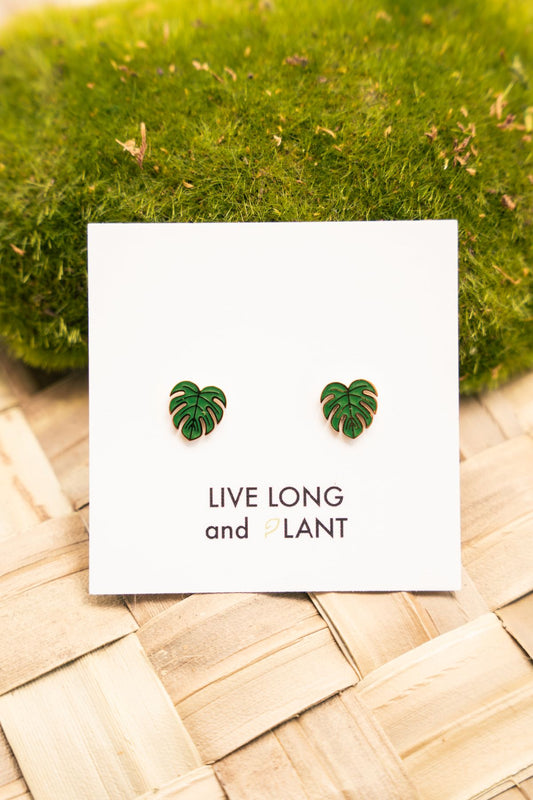A pair of green monstera leaf stud earrings displayed on a white card that reads 'LIVE LONG and PLANT', placed on a woven light wood surface with green moss in the background.