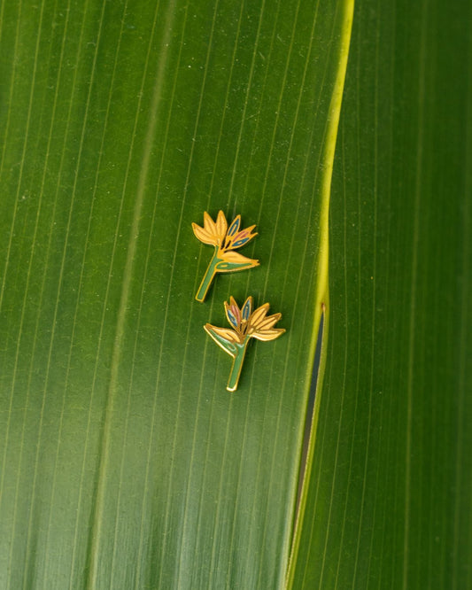 Two small gold flower-shaped earrings with green and blue accents resting on a large green leaf.