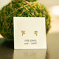 A pair of small gold flower stud earrings displayed on a white card that reads 'LIVE LONG and PLANT', set against a blurred green moss ball and tabletop background.