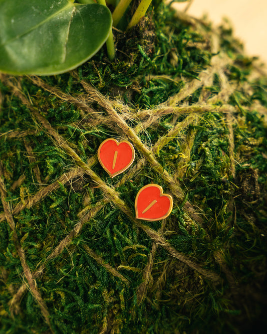 Two small red heart-shaped enamel earrings with gold borders resting on green moss wrapped in twine.