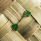 A pair of green monstera leaf stud earrings with gold-tone edges, displayed on a woven natural bamboo surface.