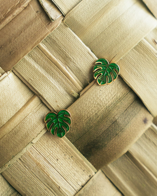 A pair of green monstera leaf stud earrings with gold-tone edges, displayed on a woven natural bamboo surface.