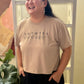 Person wearing a beige t-shirt with text, standing indoors with a plant and ladder in the background.