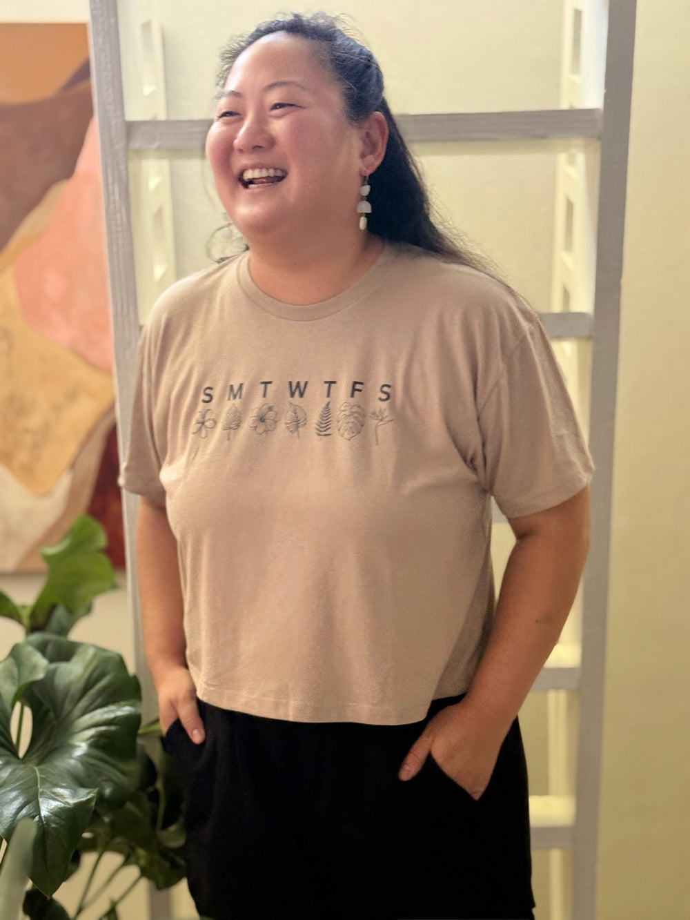 Person wearing a beige t-shirt with text, standing indoors with a plant and ladder in the background.