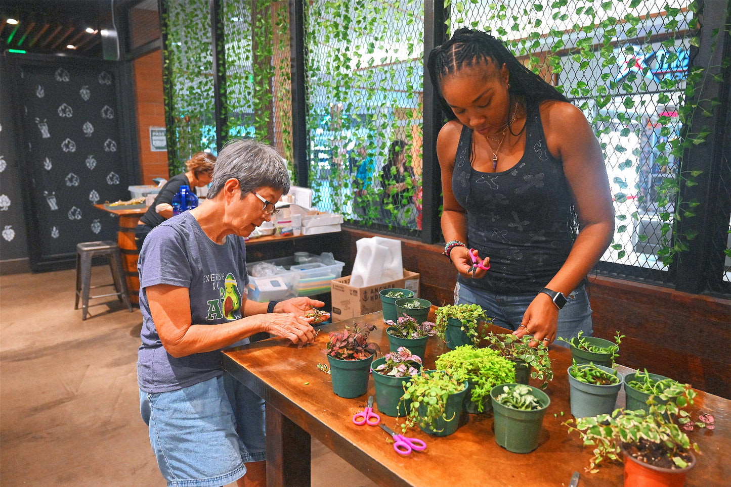 Two women working with plants at a table in a room with stained glass windows.
