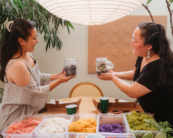 Two women holding jars with plants in a room with plants and a table.