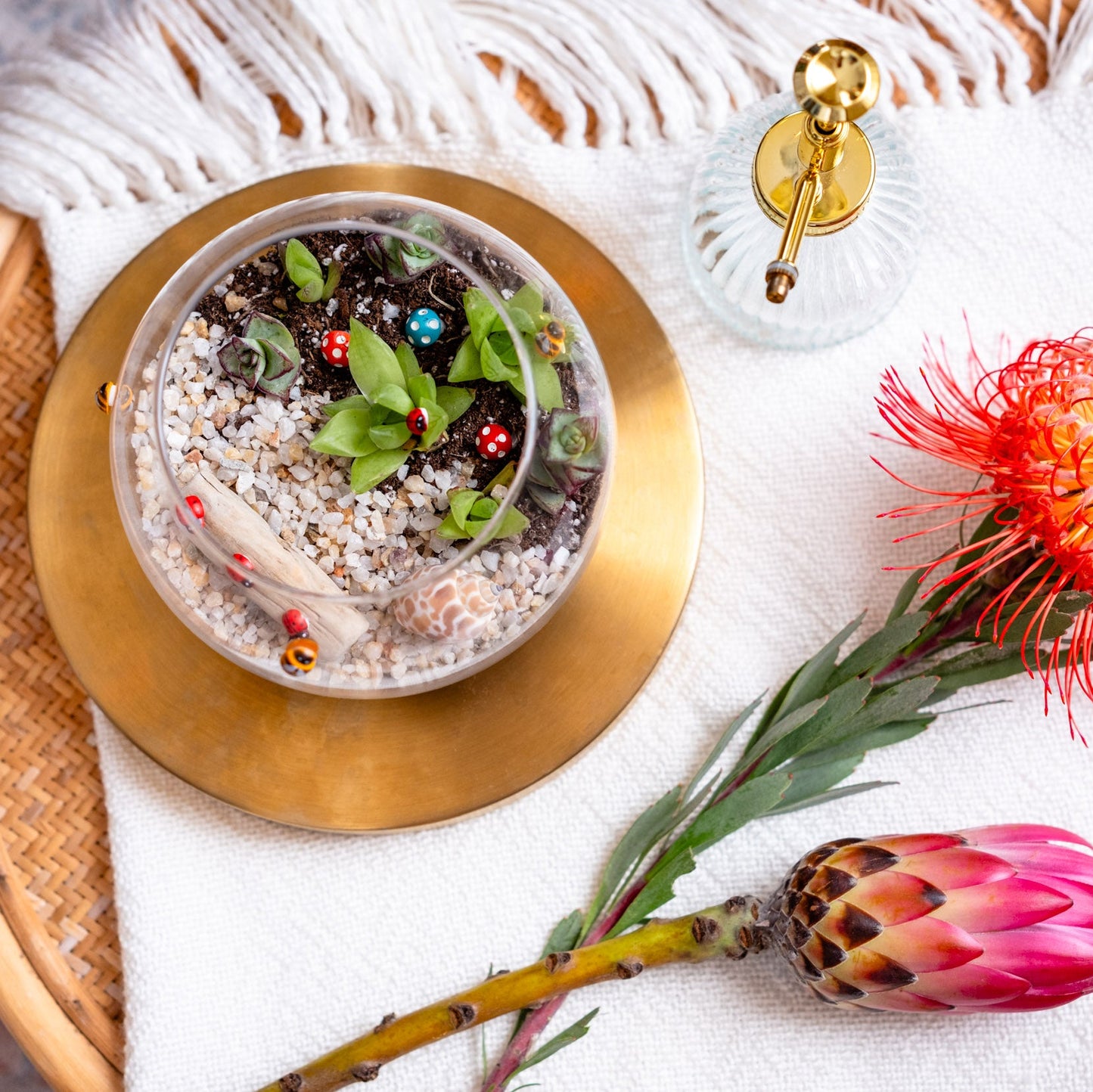 Decorative dish with sand, small plants, and colorful beads on a gold plate, accompanied by a red and pink flower on a white surface.