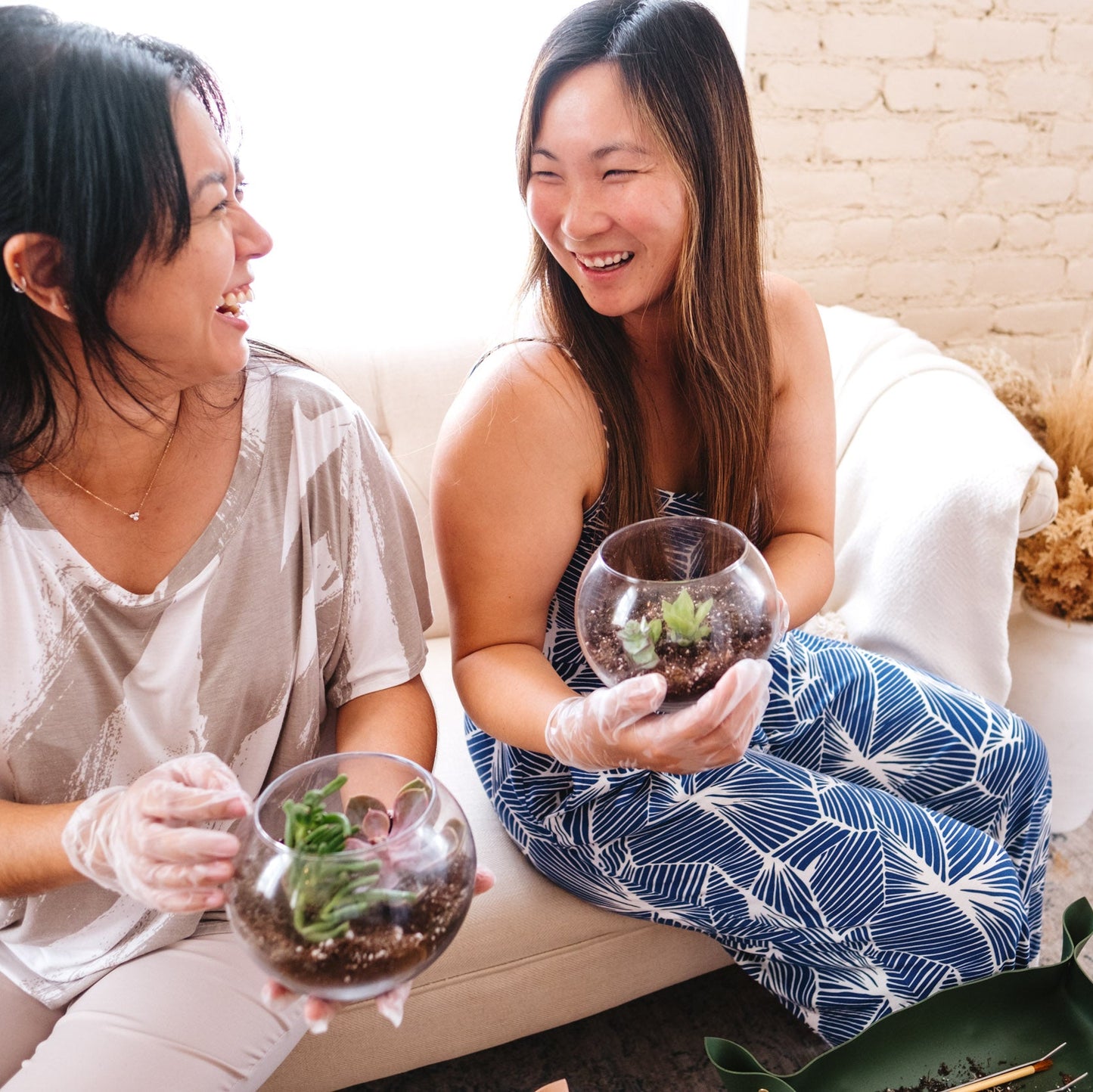 Two women sitting on a couch holding terrariums in a cozy living room.