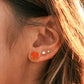 A close-up of a person's ear wearing a small orange-red heart stud earring with a gold border, alongside other ear piercings, with warm sunlight and hair visible.