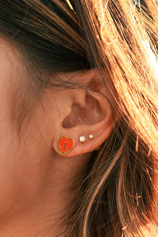 A close-up of a person's ear wearing a small orange-red heart stud earring with a gold border, alongside other ear piercings, with warm sunlight and hair visible.