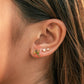 Close-up of a person's ear showcasing a stack of three small gold ear piercings: a green leaf cluster stud, a pair of tiny white pearl studs, and a tiny gold bead.