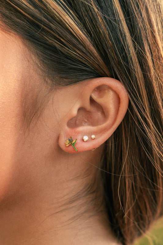 Close-up of a person's ear showcasing a stack of three small gold ear piercings: a green leaf cluster stud, a pair of tiny white pearl studs, and a tiny gold bead.