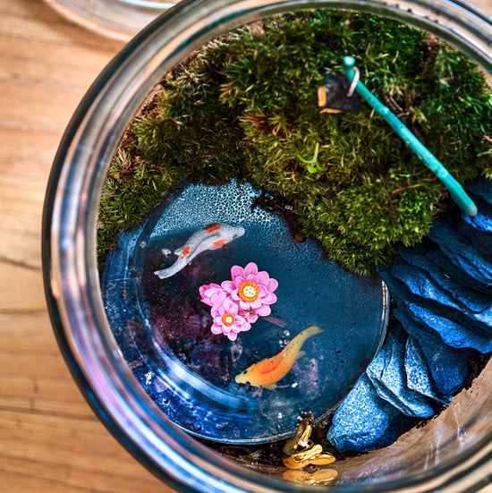 Jar with miniature pond setup including fish, flowers, and plants on a wooden surface