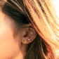A close-up of a person's ear wearing a small gold geometric stud earring and a small silver stud, with warm sunlight filtering through brown and blonde hair.