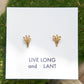 A pair of small gold stud earrings displayed on a white card that reads 'LIVE LONG and PLANT', set against a green mossy background.