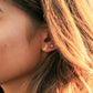 A close-up side profile of a woman's ear wearing a small gold leaf-shaped stud earring and a tiny pearl-like stud, with warm sunlight and hair visible.