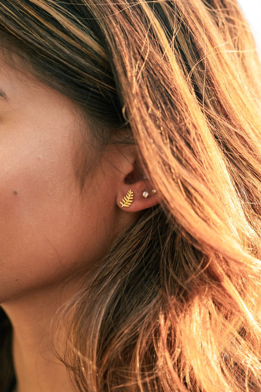 A close-up side profile of a woman's ear wearing a small gold leaf-shaped stud earring and a tiny pearl-like stud, with warm sunlight and hair visible.