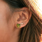 A close-up of a person's ear wearing a small green and yellow leaf stud earring, with two small pearl-like studs above it and warm sunlight reflecting off the hair.