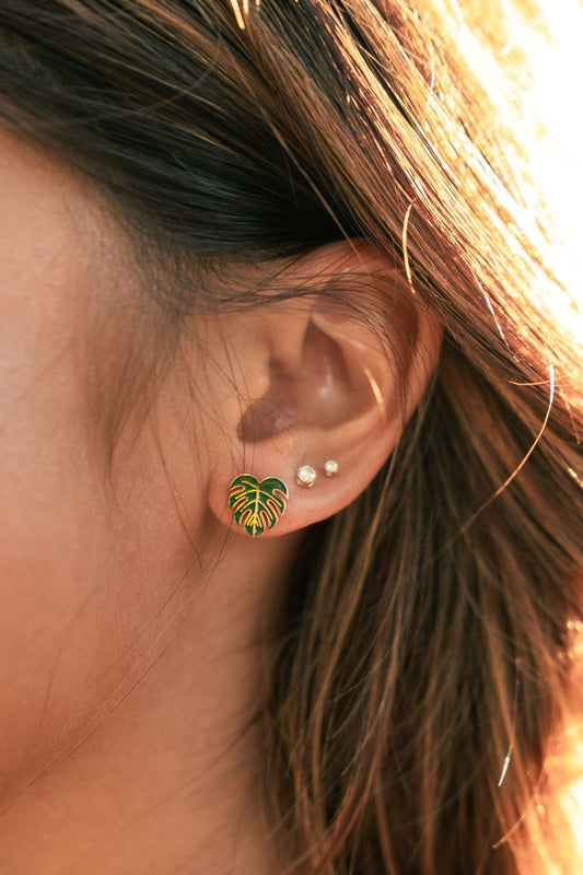 A close-up of a person's ear wearing a small green and yellow leaf stud earring, with two small pearl-like studs above it and warm sunlight reflecting off the hair.