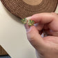 Hand holding a pair of green leaf-shaped earrings with gold accents against a woven mat background.