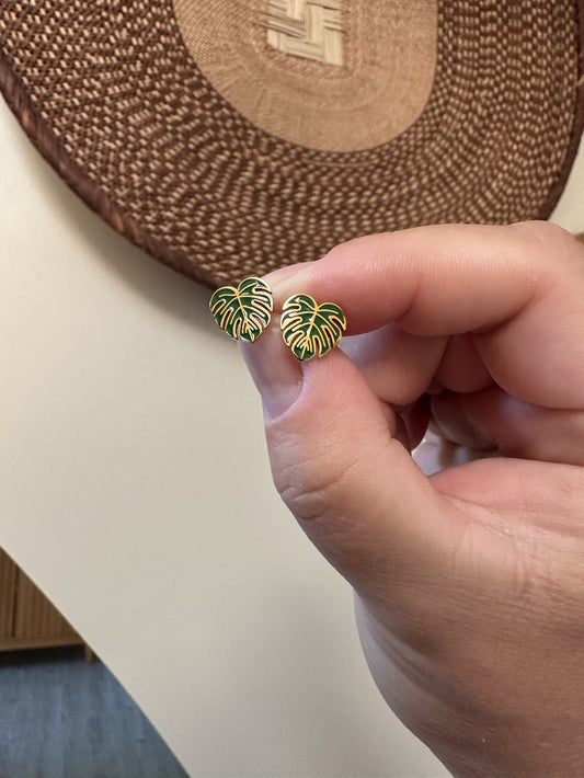 Hand holding a pair of green leaf-shaped earrings with gold accents against a woven mat background.