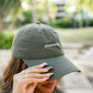 A woman outdoors wearing an olive green dad hat with the word 'plantrovert' embroidered in white on the front, holding the brim over her eye.