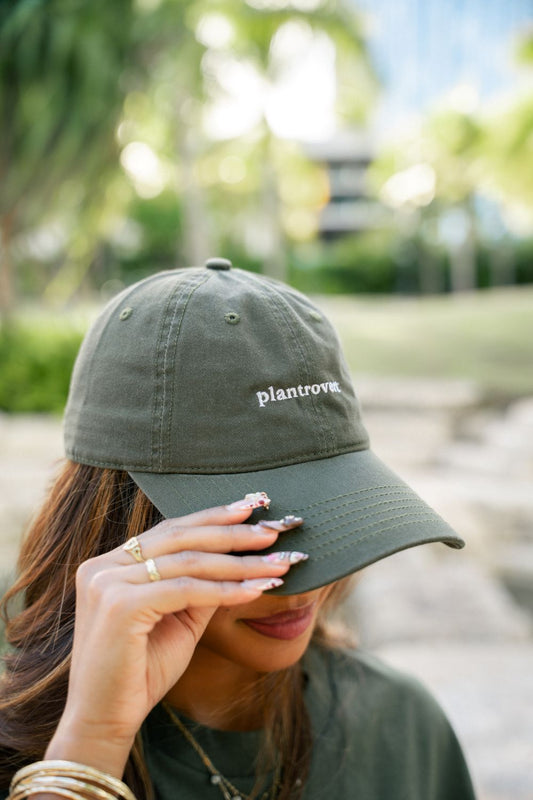 A woman outdoors wearing an olive green dad hat with the word 'plantrovert' embroidered in white on the front, holding the brim over her eye.