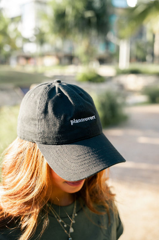 A person outdoors wearing a black baseball cap with the word "plantrovert" embroidered in white on the side. The background is a blurred park setting with trees and sunlight.
