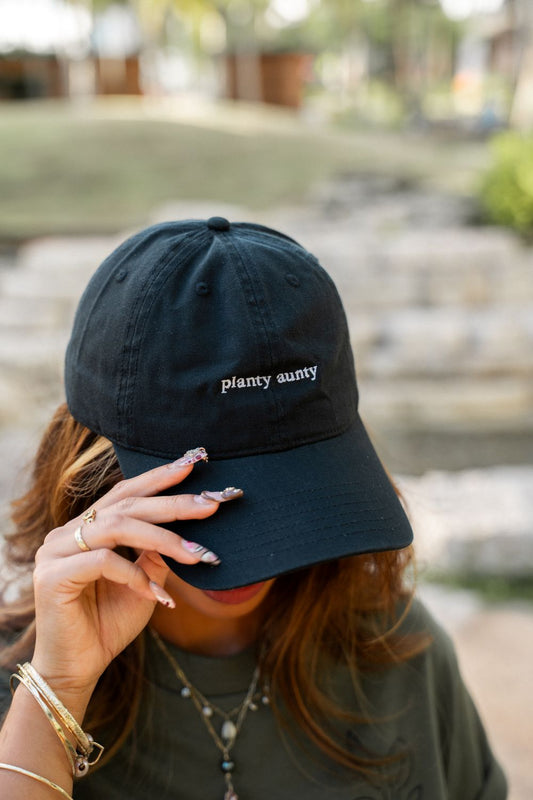 A person outdoors wearing a black baseball cap with the words 'planty aunty' embroidered in white on the front, holding the brim with one hand.